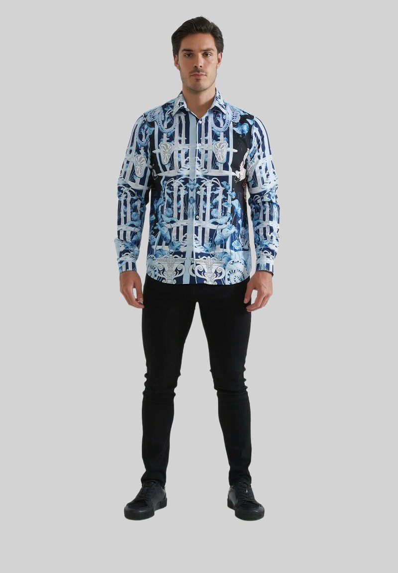 Chemise à manches longues bleu et noir, avec un motif graphique comportant des éléments architecturaux. Portée avec un pantalon slim noir et des chaussures noires.