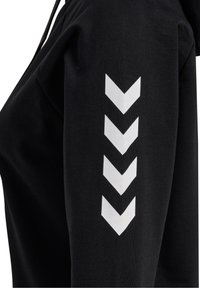 Svart sweatshirt med mjuk textur, med vita chevronmönster längs högra ärmen. Huvtröja med justerbara dragsnören.