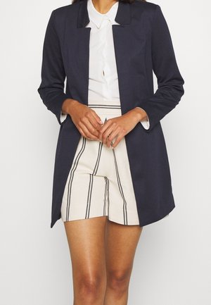 Vrouw draagt een marineblauwe blazer, een wit overhemd met knopen en crèmekleurige shorts met verticale zwarte strepen, staand met gevouwen handen voor zich.
