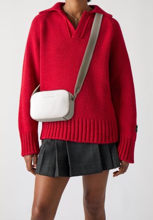 Persoon draagt een rood gebreide trui en een grijze plooirok met een witte crossbodytas met een brede grijze band.