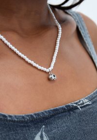 Valkoinen helminauha, jossa on hopeinen pääkallon riipus, esitelty tumman farkkukankaan taustalla. Silkkinen pintakuvio, klassinen muotoilu.