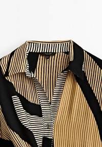 Hemdkragen und obere Brust mit gemischten vertikalen beigen und schwarzen Streifen sowie kleineren weißen und schwarzen Streifen, Knopf teilweise geschlossen.