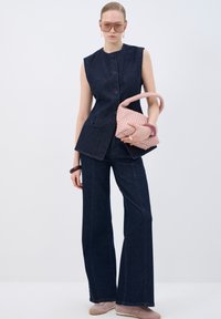 Top de denim sin mangas con cierre de botones, acompañado de unos jeans de pierna ancha oscuros. Los accesorios incluyen un bolso tejido rosa y gafas de sol extragrandes.