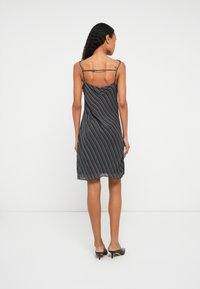 Hosszú, göndör hajú nő fekete-fehér csíkos ujjatlan ruhában és fekete magassarkú cipőben áll, háttal a kamerának, fehér háttér előtt.