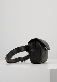 Sac en cuir noir avec une surface texturée, de forme rectangulaire et une fermeture à rabat. Sangle réglable et détail d'une poche zippée visible.