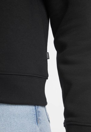 Zwarte sweatshirt met een gladde textuur, voorzien van geribbelde mouwen en zoom, en een klein logo-tag aan de zijkant. Gepaard met lichtblauwe jeans.