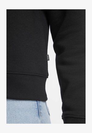 Crna dukserica glatke teksture, s rebrastim manžetama i rubom, te malim logotipom s bočne strane. Uparen s svijetloplavim trapericama.