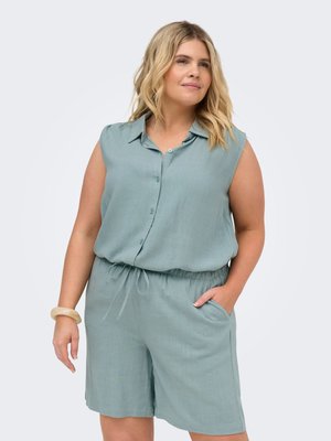 Femme aux cheveux blonds portant une chemise sans manches bleu sarcelle à boutons et un short assorti avec cordon, une main dans la poche, fond neutre.