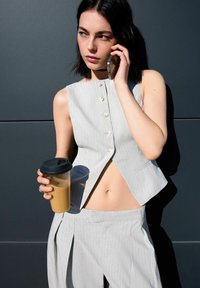 Šedý pruhovaný bezrukávový crop top s gombíkmi, v kombinácii s vysooko pasovými nohavicami. Modelka drží priehľadný pohár s čiernym viečkom.