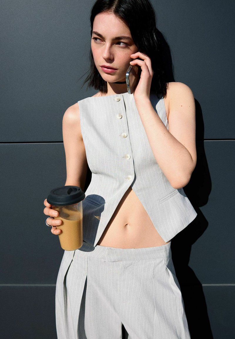 Šedý pruhovaný bezrukávový crop top s gombíkmi, v kombinácii s vysooko pasovými nohavicami. Modelka drží priehľadný pohár s čiernym viečkom.