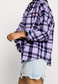 Fialová kostkovaná flanelová košile s černými akcenty a malou nášivkou s logem. Materiál vypadá měkce, má volný střih a zapínání na knoflíky.