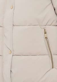 Veste rembourrée beige clair avec un matelassage en losanges, dotée d'une poche zippée sur le côté avec des garnitures dorées et de fermetures à pression sur le devant.