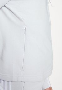 Gros plan sur un sweat à capuche gris clair montrant une poche latérale verticale zippée et des détails en tissu texturé.