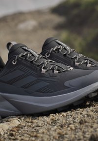 Chaussures de trail Adidas Terrex noires et grises reposant sur du gravier, avec des lacets réfléchissants et des semelles texturées, pour une utilisation en extérieur.