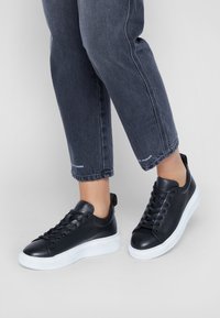 Baskets en cuir noir à texture lisse, bout rond, semelles en caoutchouc blanc et lacets plats. Portées avec un pantalon en denim foncé.