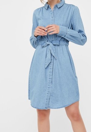 Kobieta ubrana w jasnoniebieską sukienkę-koszulę z długimi rękawami, kołnierzykiem, guzikami z przodu oraz paskiem zawiązywanym w talii.