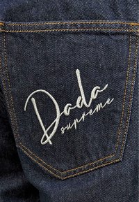 Dark blue denim fabric with visible texture, featuring an embroidered white logo "Dada supreme" on the back pocket, outlined in orange stitching.
