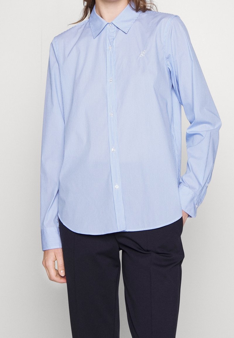 Femme portant une chemise boutonnée bleu clair à fines rayures rentrée dans un pantalon noir, debout avec une main dans la poche sur fond blanc.