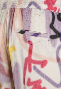 Nahaufnahme eines Stoffes mit Graffiti-Stil Mustern in Rot, Lila, Gelb und Schwarz, die mit Sprühfarben aufgetragen sind, sowie einer kleinen horizontalen Taschenöffnung.