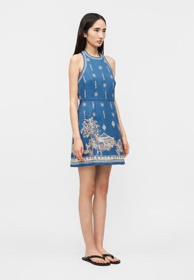 Γυναίκα που στέκεται με μπλε αμάνικο φόρεμα με λεπτομερή μπεζ σχέδιο με φοίνικες και σκηνές αγοράς, φοράει μαύρες σαγιονάρες σε λευκό φόντο.
