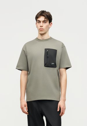 Νέος άνδρας φοράει υπερμεγέθη ελιάς πράσινη μπλούζα με μεγάλη μαύρη τσέπη στο στήθος με φερμουάρ και μαύρο παντελόνι, στέκεται απέναντι σε απλό φωτεινό φόντο.