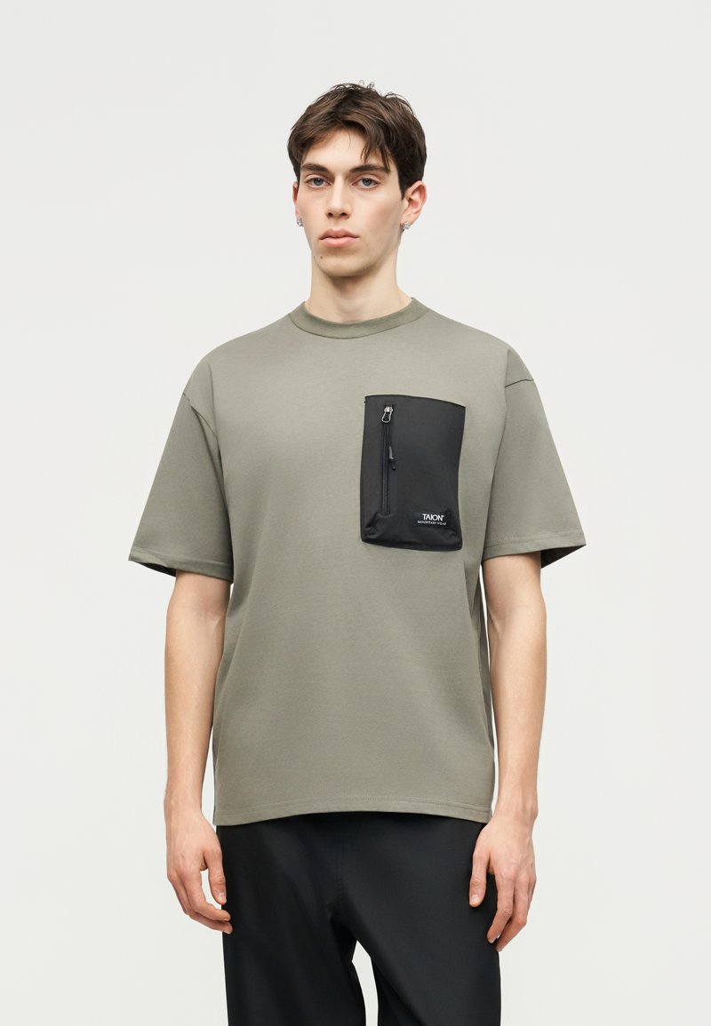 Νέος άνδρας φοράει υπερμεγέθη ελιάς πράσινη μπλούζα με μεγάλη μαύρη τσέπη στο στήθος με φερμουάρ και μαύρο παντελόνι, στέκεται απέναντι σε απλό φωτεινό φόντο.