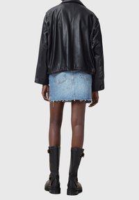 Personne portant une veste en cuir noire, une mini-jupe en jean bleu clair effilochée et des bottes noires à boucle, debout de dos sur un fond uni.