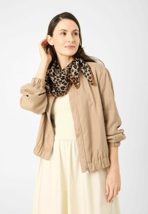 Woman wearing beige jacket over cream dress with leopard print scarf, touching hair, looking to the side against plain background.