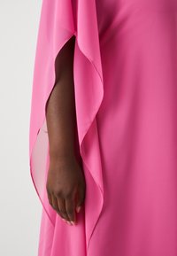 Un bras à la peau foncée visible à travers la fente d'un vêtement fluide rose vif sur un fond clair et uni.