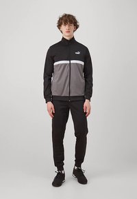 Mladić stoji odjeven u crno-sivi Puma trenerku s crnim teniskama, protiv jednostavne svijetlosive pozadine.