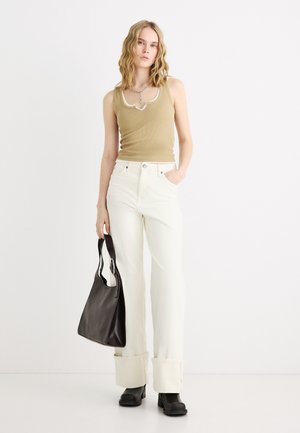 Femme aux cheveux blonds ondulés portant un haut beige sans manches, un pantalon blanc taille haute à jambes larges, des bottes noires, et tenant un sac à main noir.