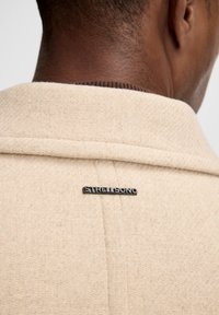 Collo di giacca beige con texture, con un'etichetta logo, caratterizzato da un sottile motivo a spina di pesce e una finitura liscia lungo il collo.