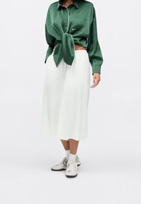 Personne portant une chemise en satin vert foncé boutonnée et nouée à la taille, une jupe midi plissée blanche, des chaussettes blanches et des baskets Nike grises.