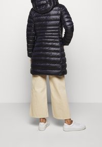 Lang dunjakke med horisontale quiltede mønster, mørk navy farve, hætte og skinnende stof. Kombination med cremede bredde bukser og hvide sneakers.