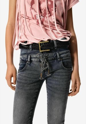 Gros plan sur une personne portant un jean noir délavé avec un lacet devant et une ceinture tressée noire avec une boucle en laiton, associé à un t-shirt rose à imprimé.