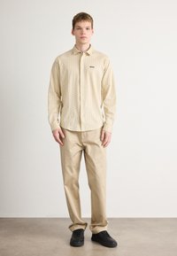 Lichtbeige gemusterd overhemd met lange mouwen en knoopsluiting, voorzien van een klein logo, gecombineerd met beige broek en zwarte sneakers op een neutrale achtergrond.
