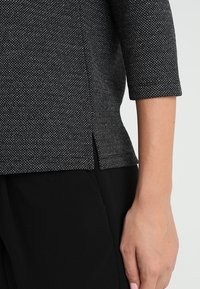 Top texturizado en blanco y negro con mangas de tres cuartos, que presenta aberturas laterales, combinado con pantalones negros. Tejido suave con un patrón sutil.