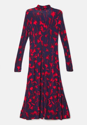 Femme portant une robe bleu marine à imprimé floral rouge, des bottines noires à bout pointu et talons, avec un sac bandoulière bleu, posant devant un mur gris uni.