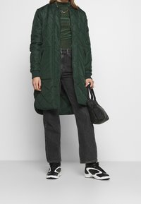 Dunkelgrüner, gesteppten langer Mantel mit gerippten Bündchen, kombiniert mit einem grün gestreiften Oberteil und schwarzen, weit geschnittenen Jeans, abgerundet mit schwarz-weißen Sneakers.