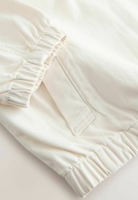 Približen posnetek belih hlač iz blaga z elastičnimi manšetami in našitim žepom z zaponko na strani.