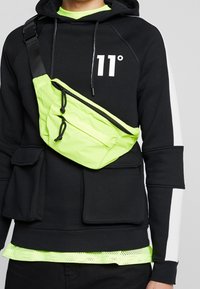 Černá mikina s kapucí s bílým logem "11°", doplněná neonově zelenou kabelkou přes rameno a viditelným spodním lemem síťovaného trička ve stejném odstínu pod mikinou.