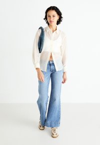 Camisa blanca transparente de botones con un corte relajado, combinada con jeans acampanados de color azul claro y zapatillas beiges, con una bolsa de denim al hombro.