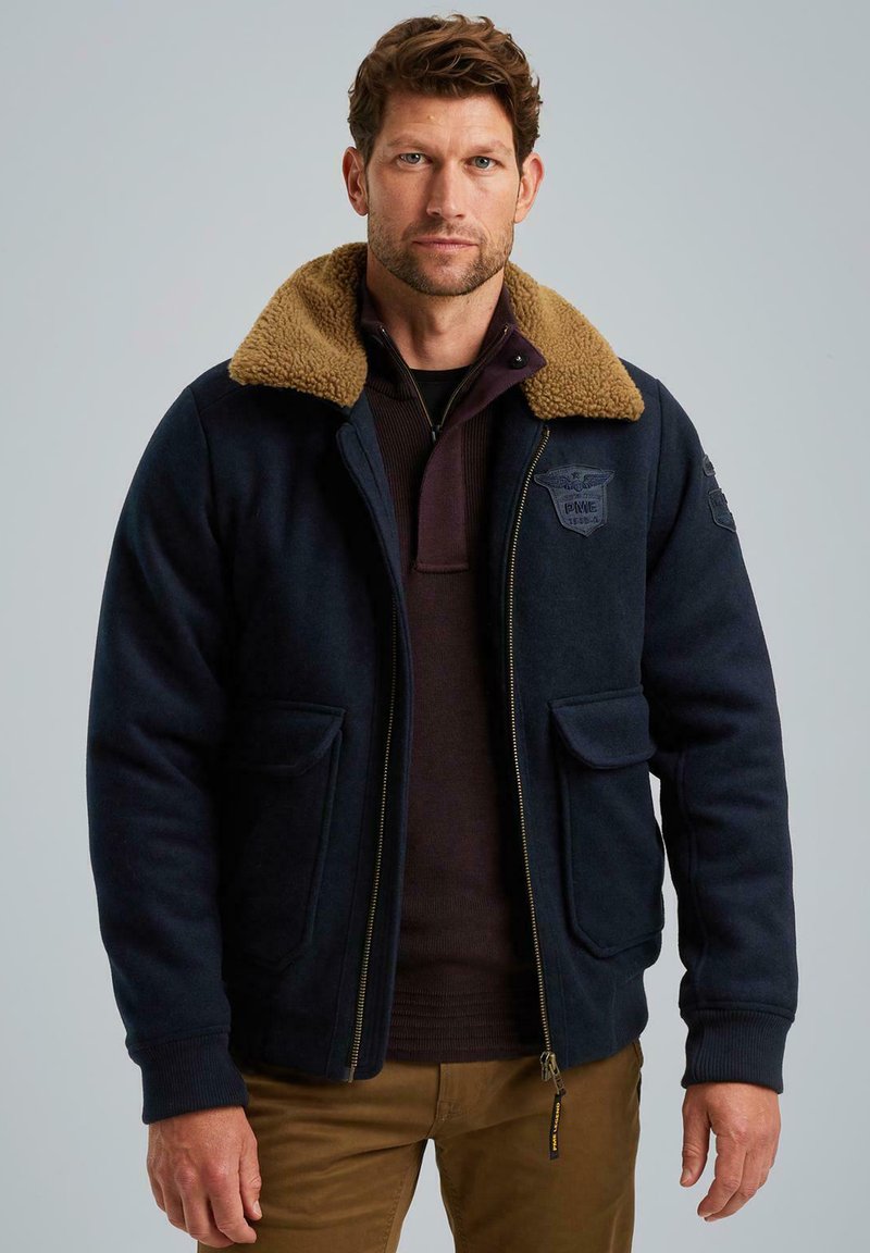 Námořnická modrá bunda s béžovým kožešinovým límcem, zipovým zapínáním, dvěma předními kapsami a logem na hrudi. Měkká textura a pevný design.
