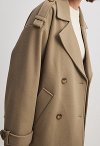 Beżowy płaszcz wełniany o dwurzędowym kroju, z dużymi guzikami, detalami naramienników na ramionach oraz bocznymi kieszeniami. Miękka faktura i zdefiniowana sylwetka.
