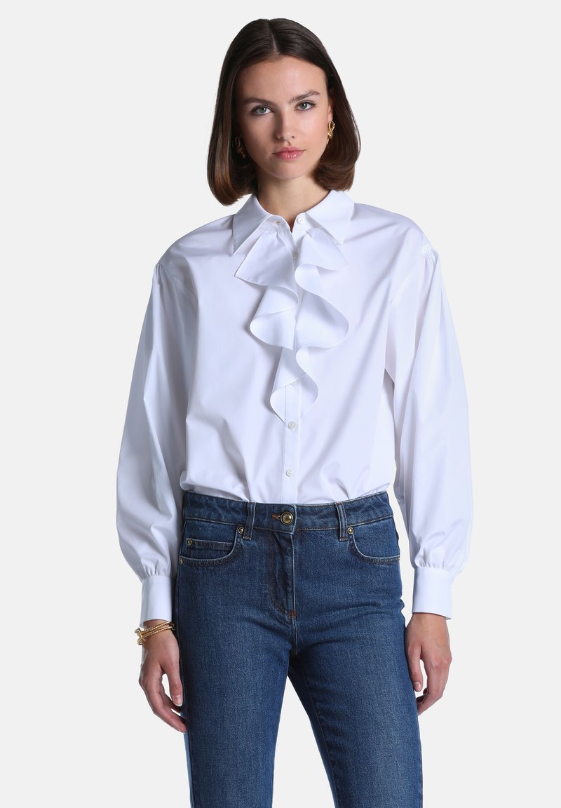 Femme portant un chemisier blanc à manches longues avec des volants sur le devant et un jean bleu en denim, debout devant un fond clair uni.