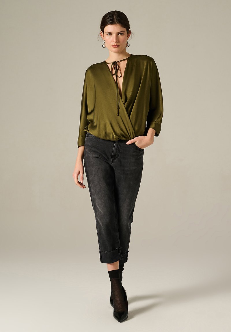 Blouse en satin vert olive avec un décolleté en V plongeant et un détail à nouer, associée à un jean foncé et des escarpins noirs à bout pointu.