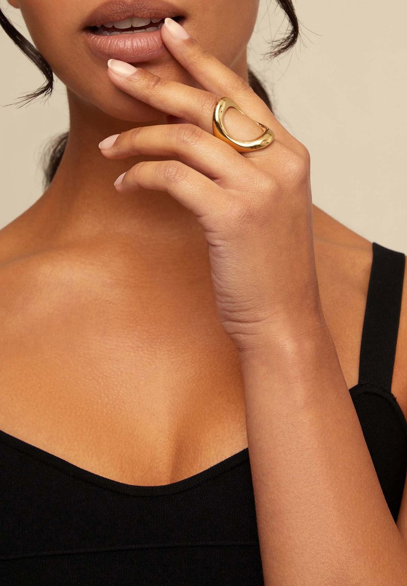 Anillo de oro con un diseño recortado que se lleva en una mano. La mano tiene las uñas bien arregladas y la persona lleva una blusa negra.