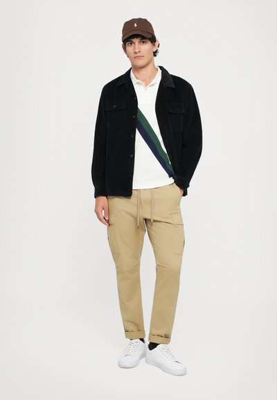 Black corduroy jacket, white polo with green and blue stripes, beige cargo pants, white sneakers, and a brown cap with a logo.