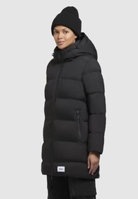 Jachetă căptușită neagră cu glugă, secțiuni mari matlasate, buzunare laterale cu fermoar și o etichetă cu logo la tiv. Purtați cu o căciulă tricotată neagră.