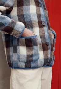 Kurtka polarowa w kratę w kolorach niebieskim, brązowym i kremowym, z guzikami z przodu oraz bocznymi kieszeniami wykończonymi niebieską lamówką.